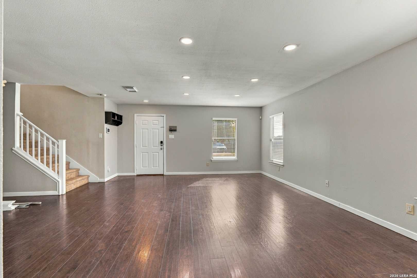 Empty living room with dark wood floors, front door, and carpeted staircase.