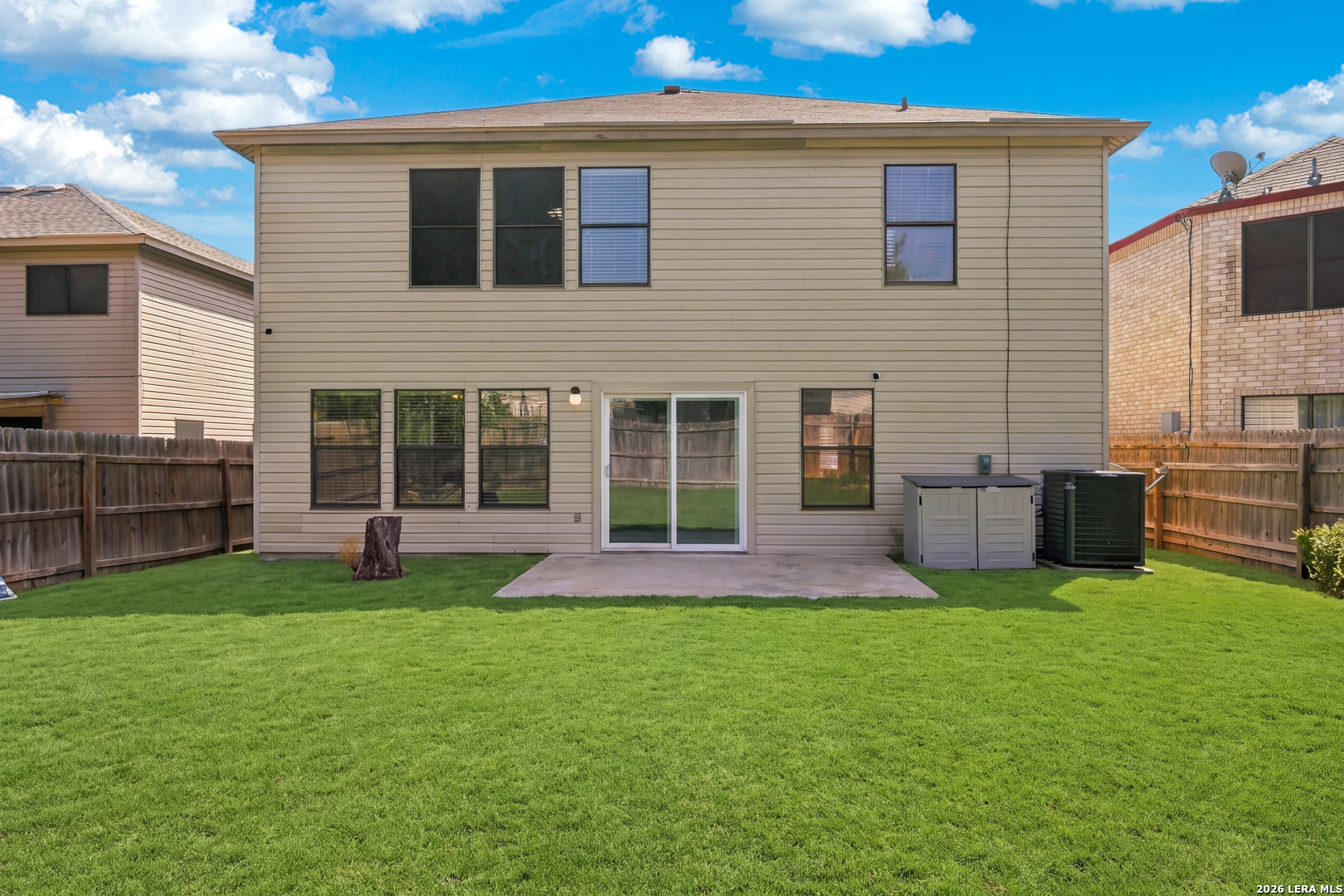 Rear of a two-story beige home with a fenced lawn, concrete patio, and sliding glass doors.