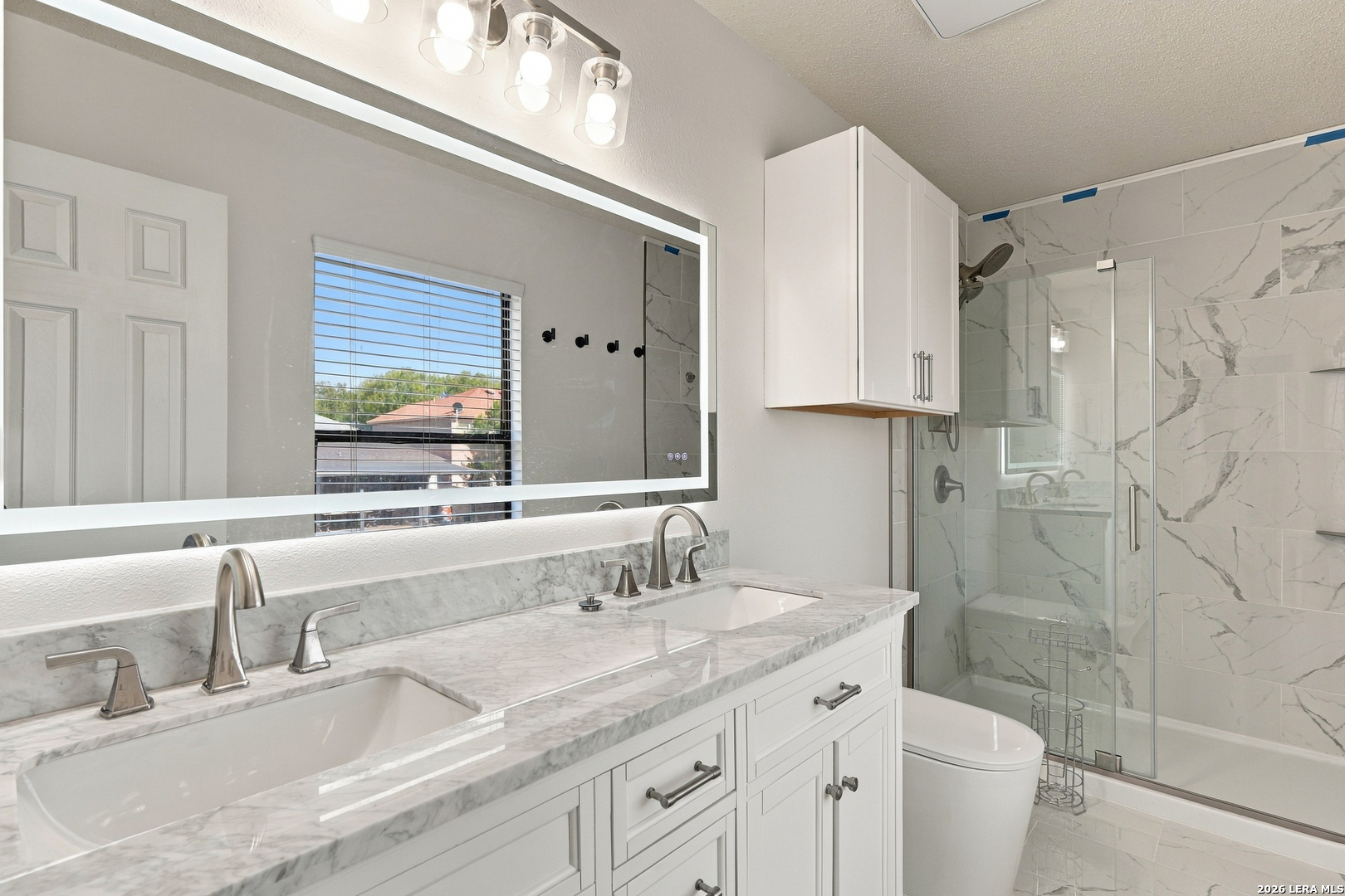 Bathroom with a double vanity and marble-look finishes beside a glass-enclosed shower.