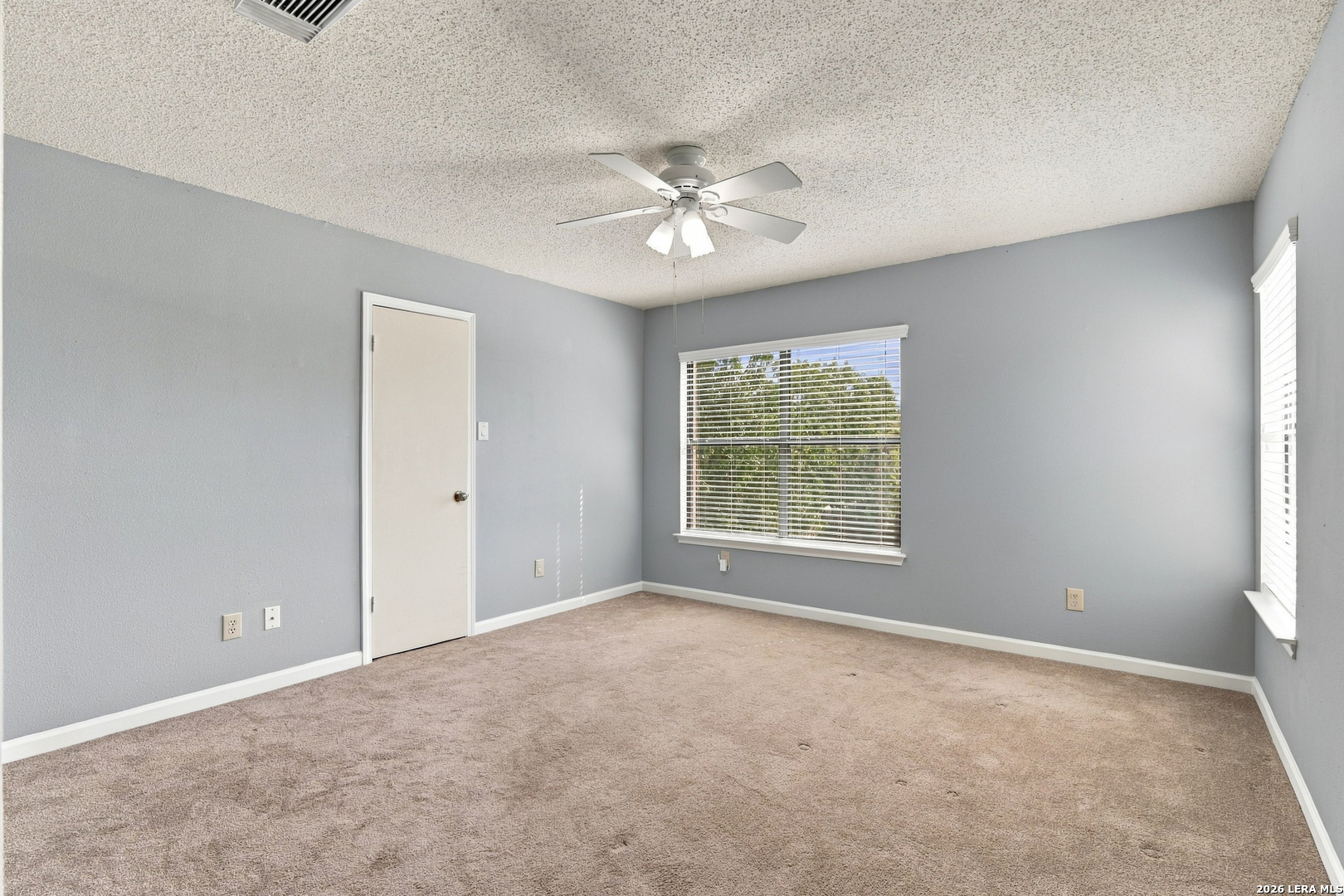 Empty bedroom with beige carpet, gray walls, ceiling fan, and two windows with blinds.