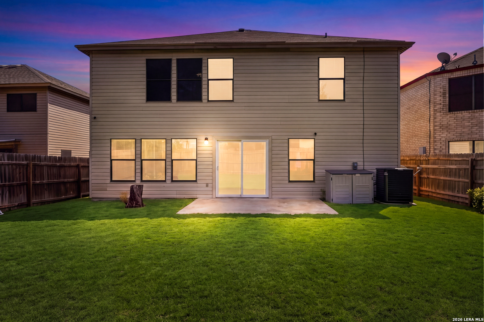 Two-story home’s rear exterior at dusk, with a broad lawn, concrete patio, and glowing windows.