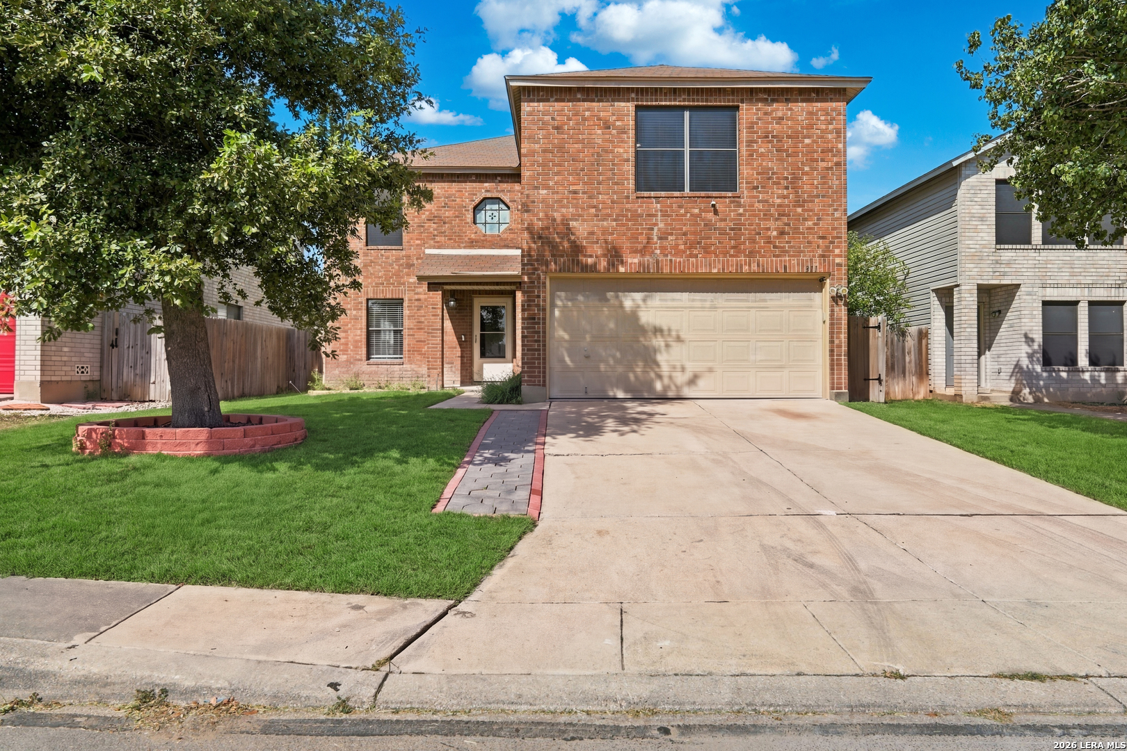 Two-story red-brick house with a beige garage, broad driveway, and a large shade tree in the front lawn.