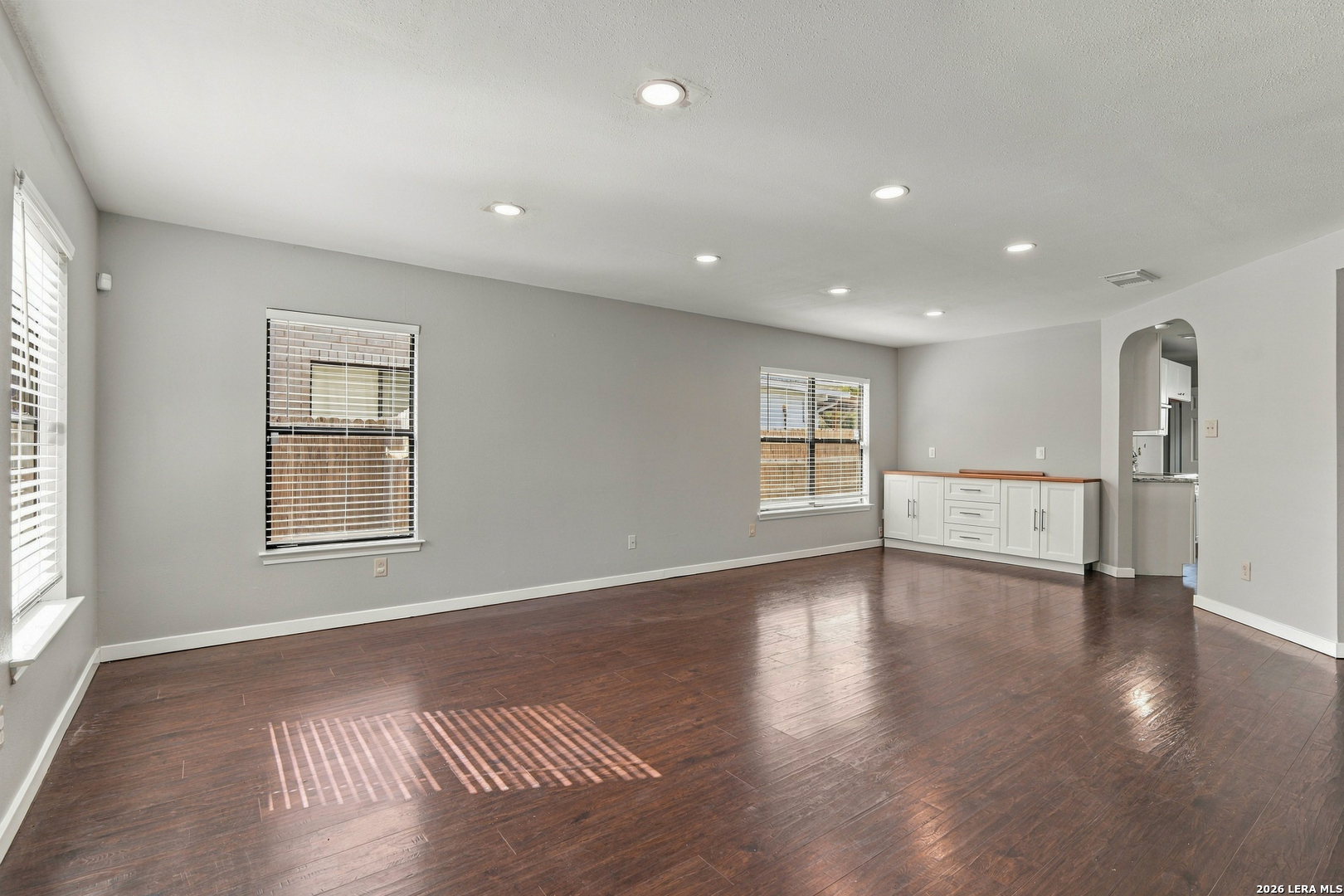 Empty living room with dark wood floors, bright windows, and a white built-in cabinet.