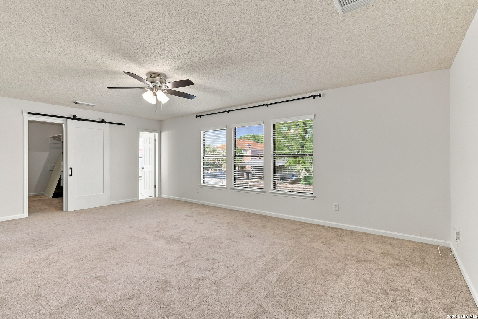 Vacant bedroom with beige carpet, a ceiling fan, three windows, and a sliding closet door.