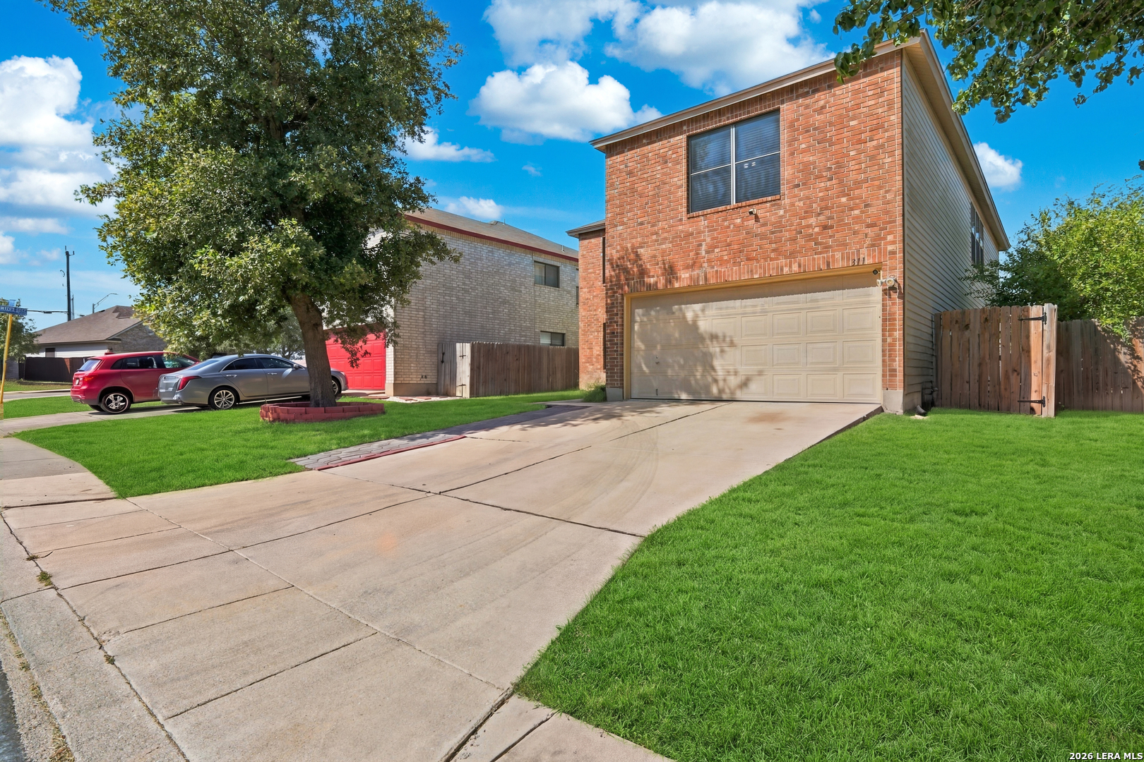 Two-story brick house with a double garage, wide driveway, green lawn, and mature shade tree.