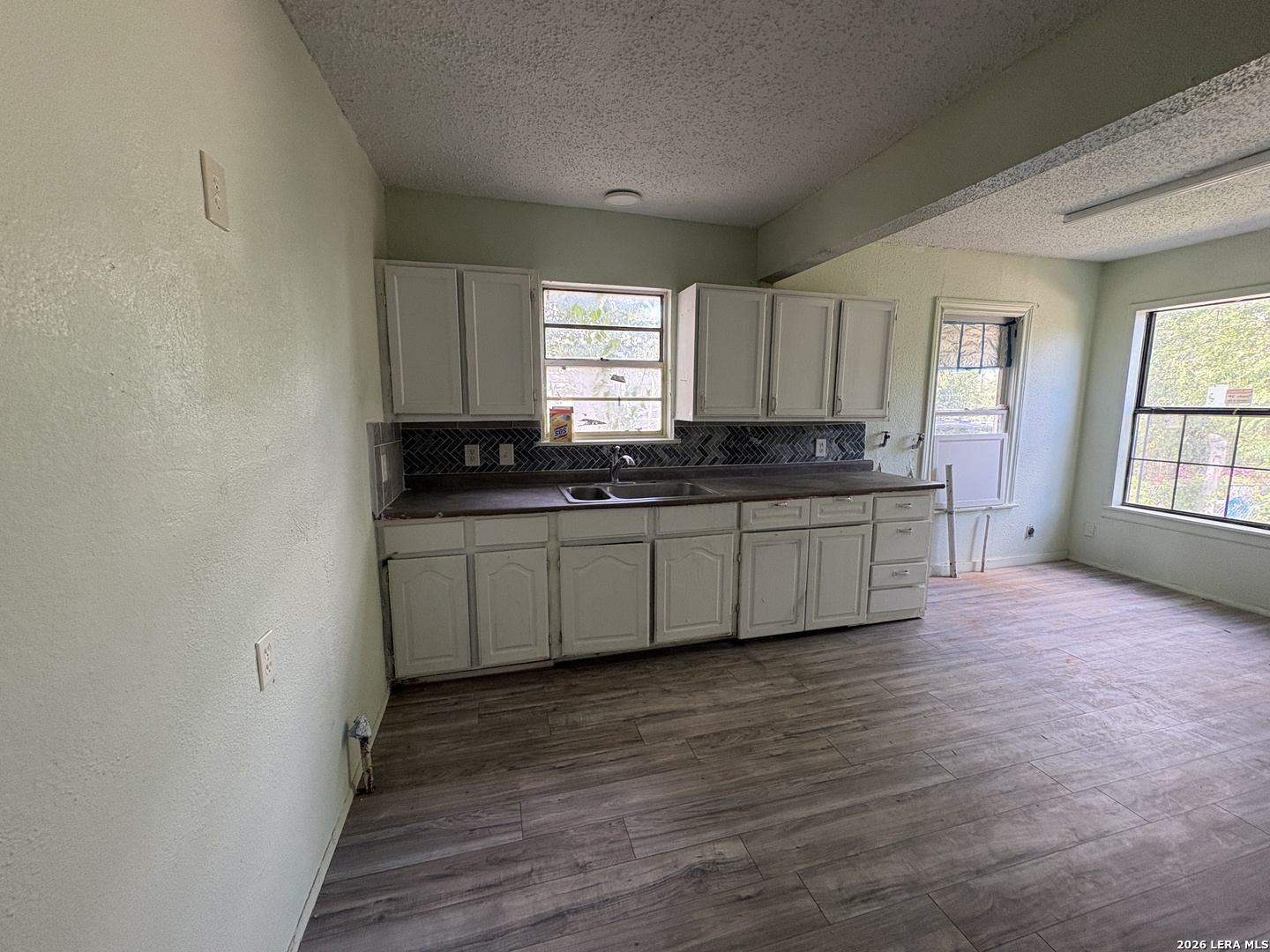 Vacant kitchen with white cabinets, a double sink, dark backsplash, and wood-look flooring.