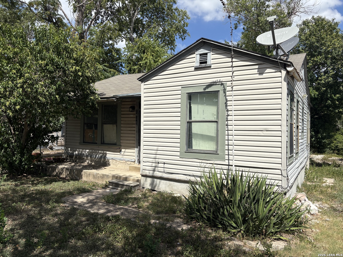 Small light-sided house with green-trimmed windows, a satellite dish, and a shaded front yard.