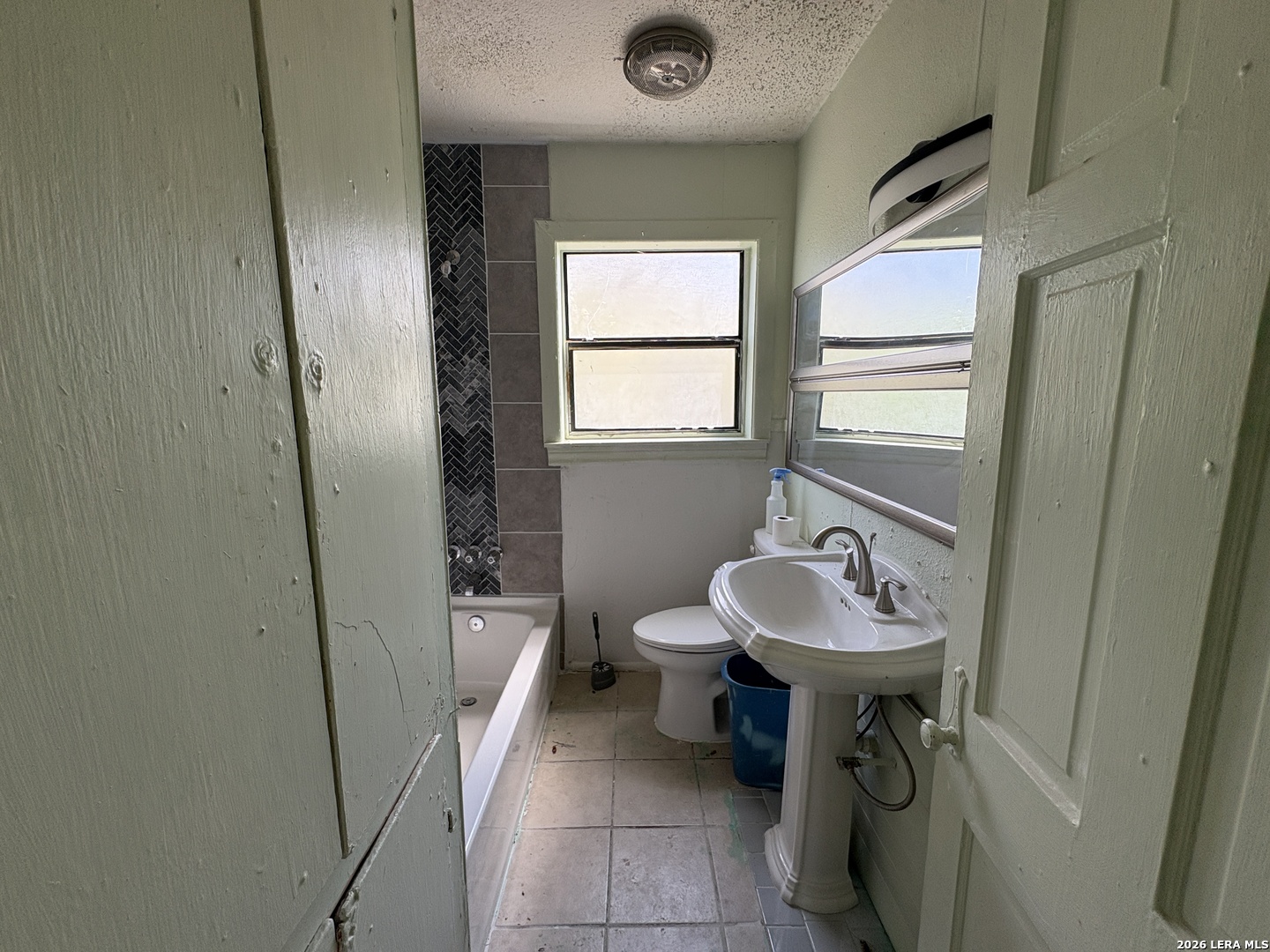 Bathroom with a bathtub, dark tiled shower wall, pedestal sink, and two windows.