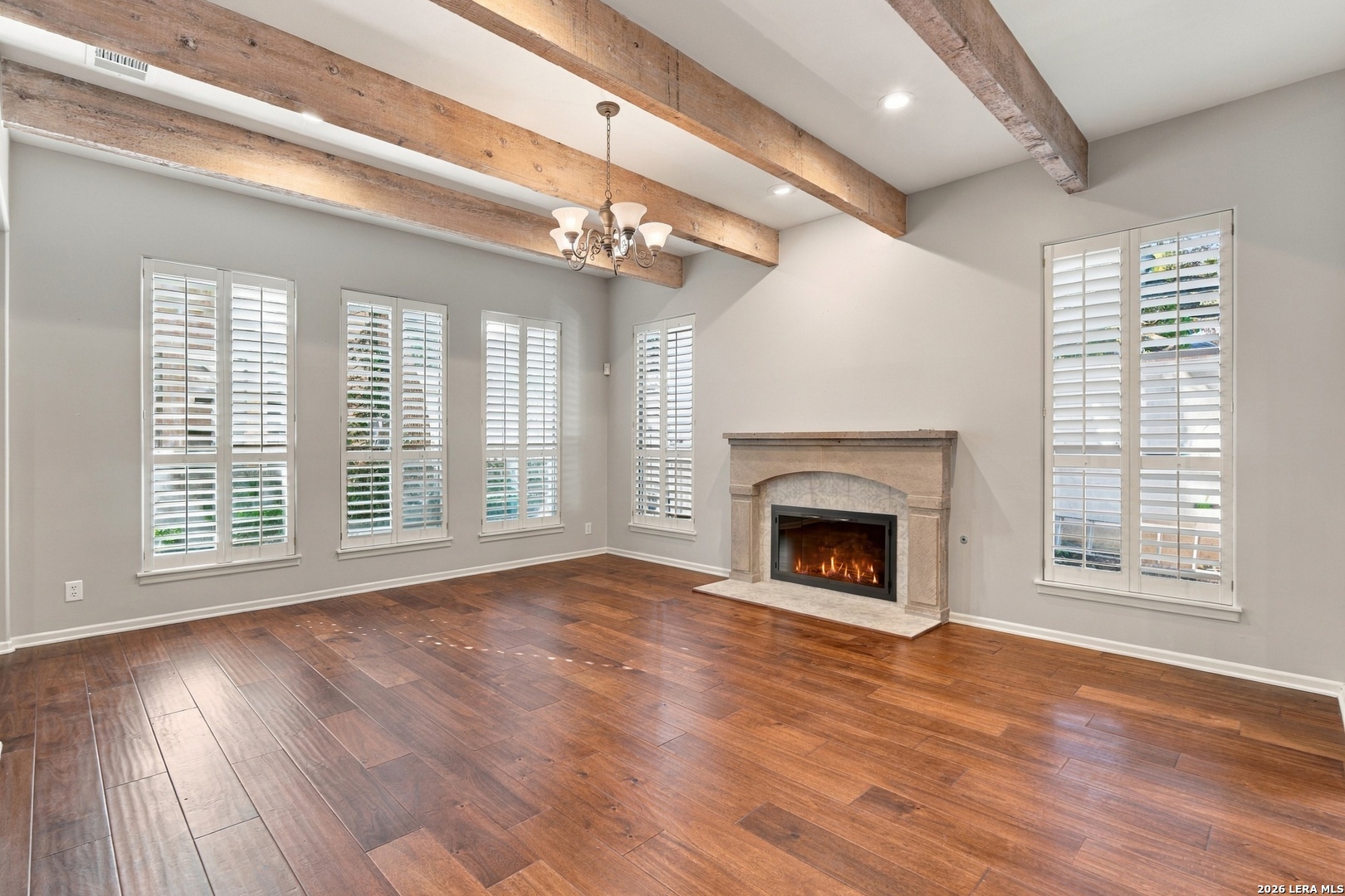 Unfurnished living room with exposed wood beams, hardwood floors, and a lit stone fireplace.