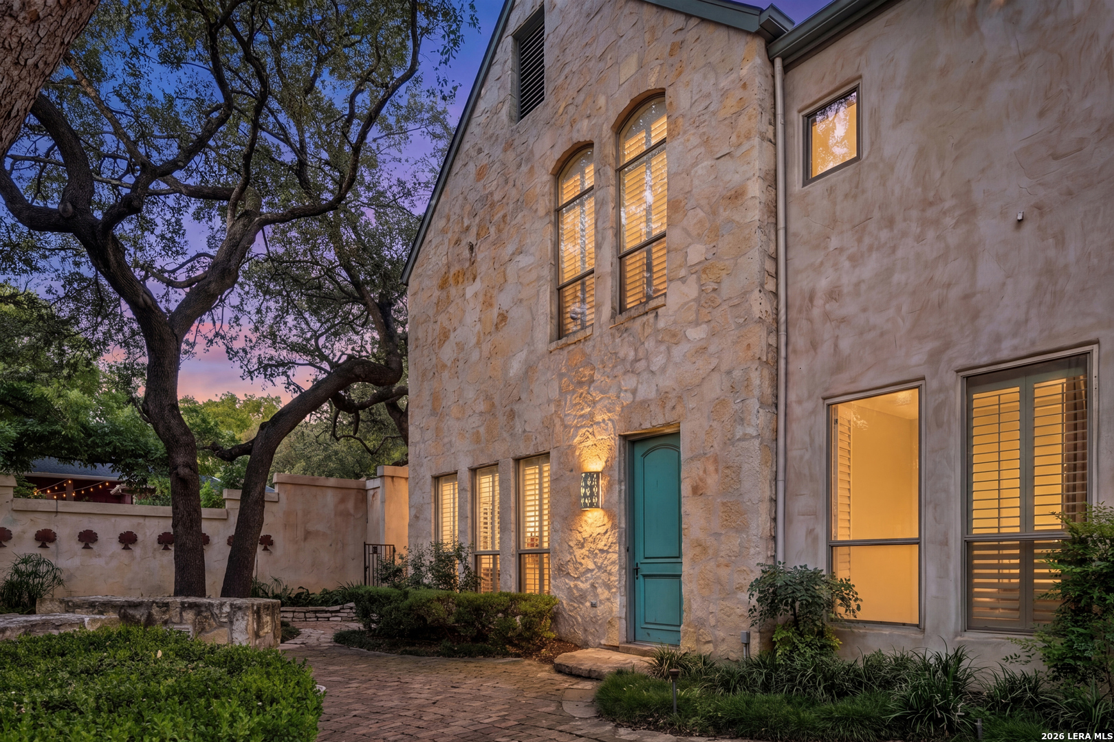 Stone-and-stucco home with a turquoise door and oak-shaded courtyard at dusk.