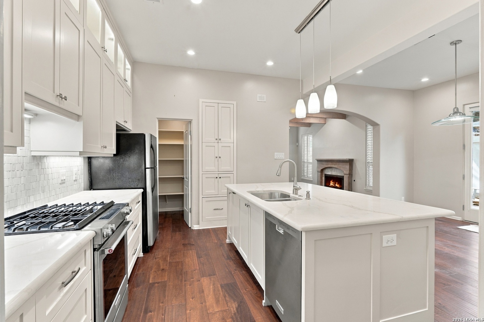 Open kitchen with a large white island, stainless appliances, dark wood floors, and an arched view of a fireplace.