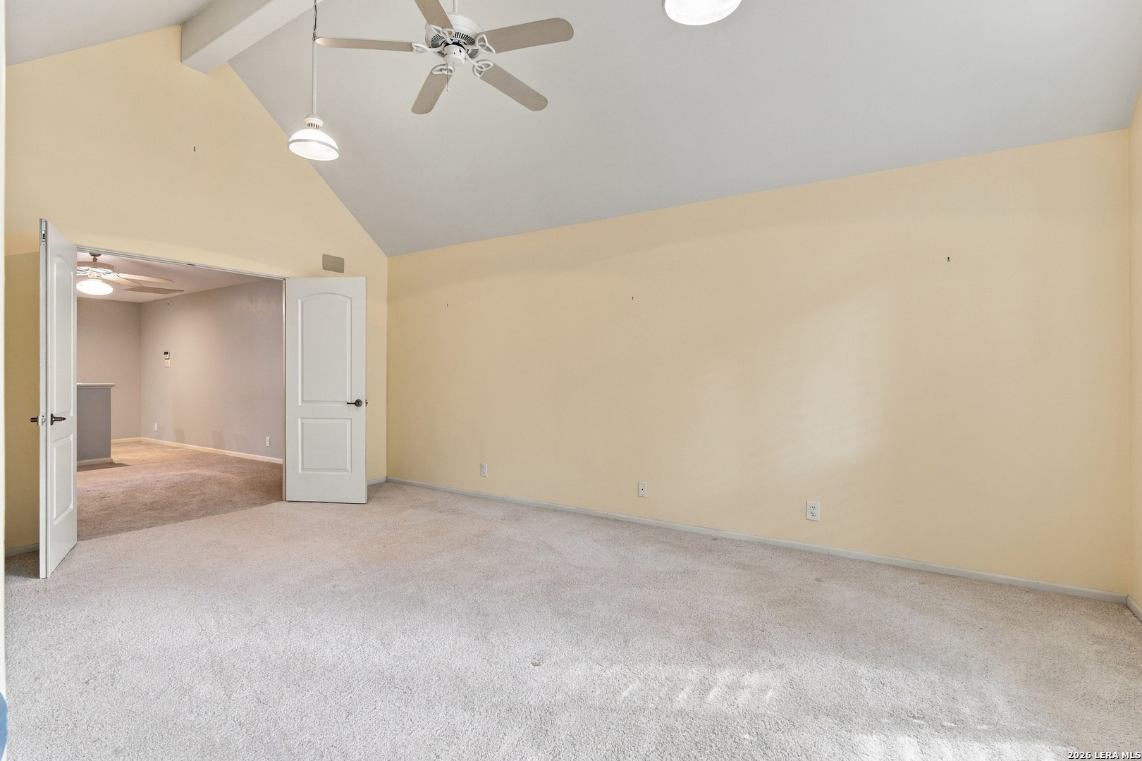 An empty carpeted room with a vaulted ceiling, ceiling fan, and open double doors.