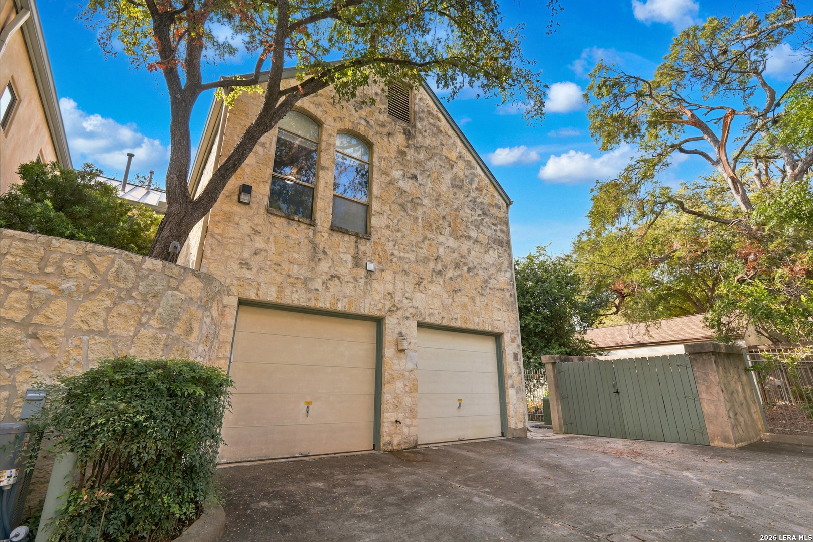 Two-story stone house with twin garage doors, driveway, mature trees, and a wooden gate.