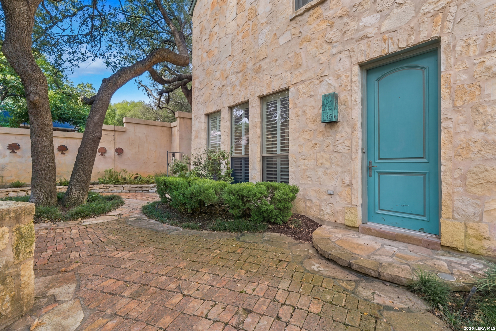 Stone home entrance with a turquoise door, brick courtyard path, and mature trees.