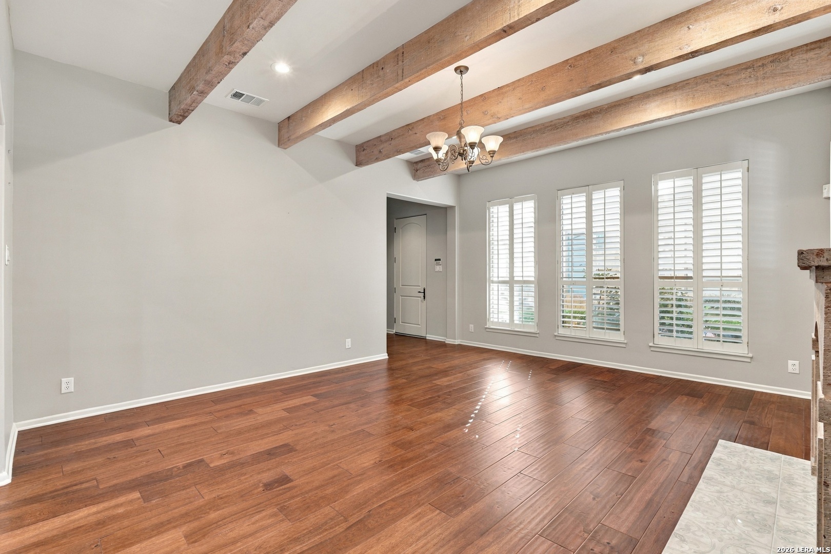 Vacant living room with exposed wood beams, hardwood floors, and tall shuttered windows.
