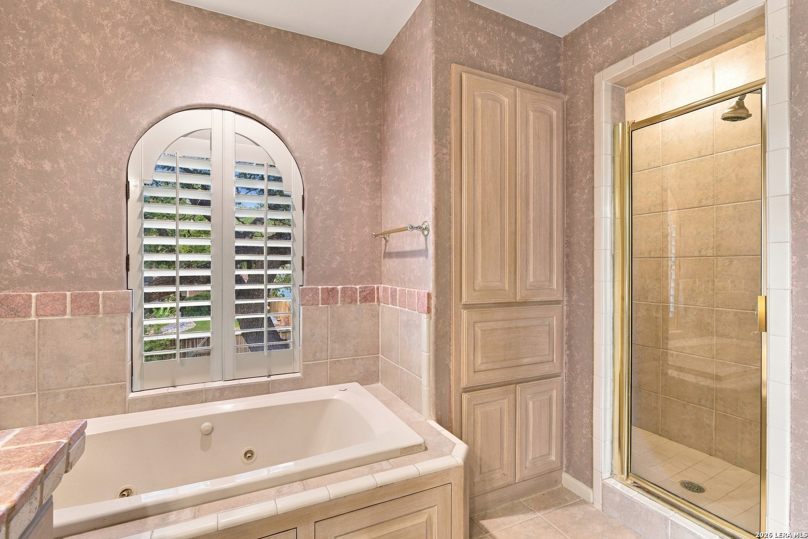 Bathroom with a built-in soaking tub, arched shuttered window, and glass-enclosed tiled shower.