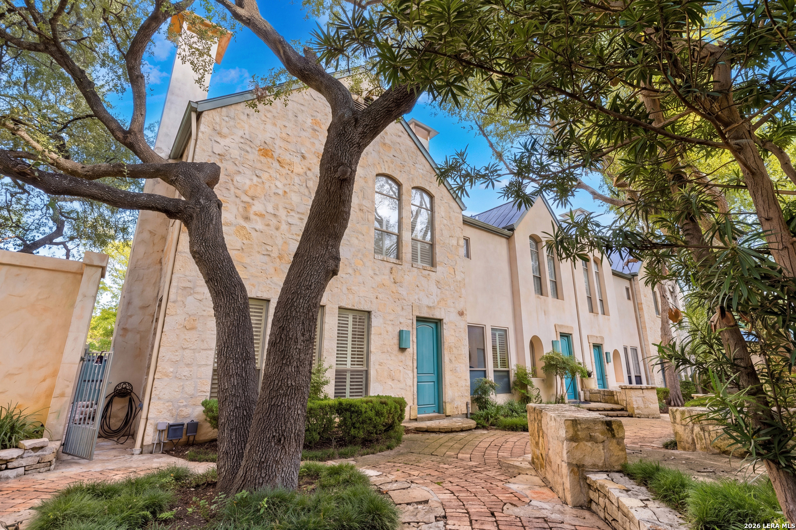 Attached stone and stucco homes with turquoise doors along a tree-shaded brick courtyard.