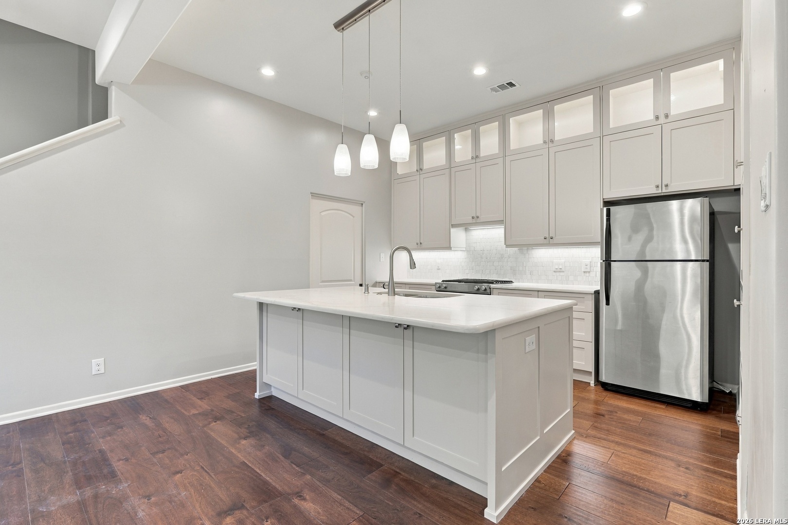 Modern white kitchen with a large island, pendant lights, stainless steel refrigerator, and dark wood floors.