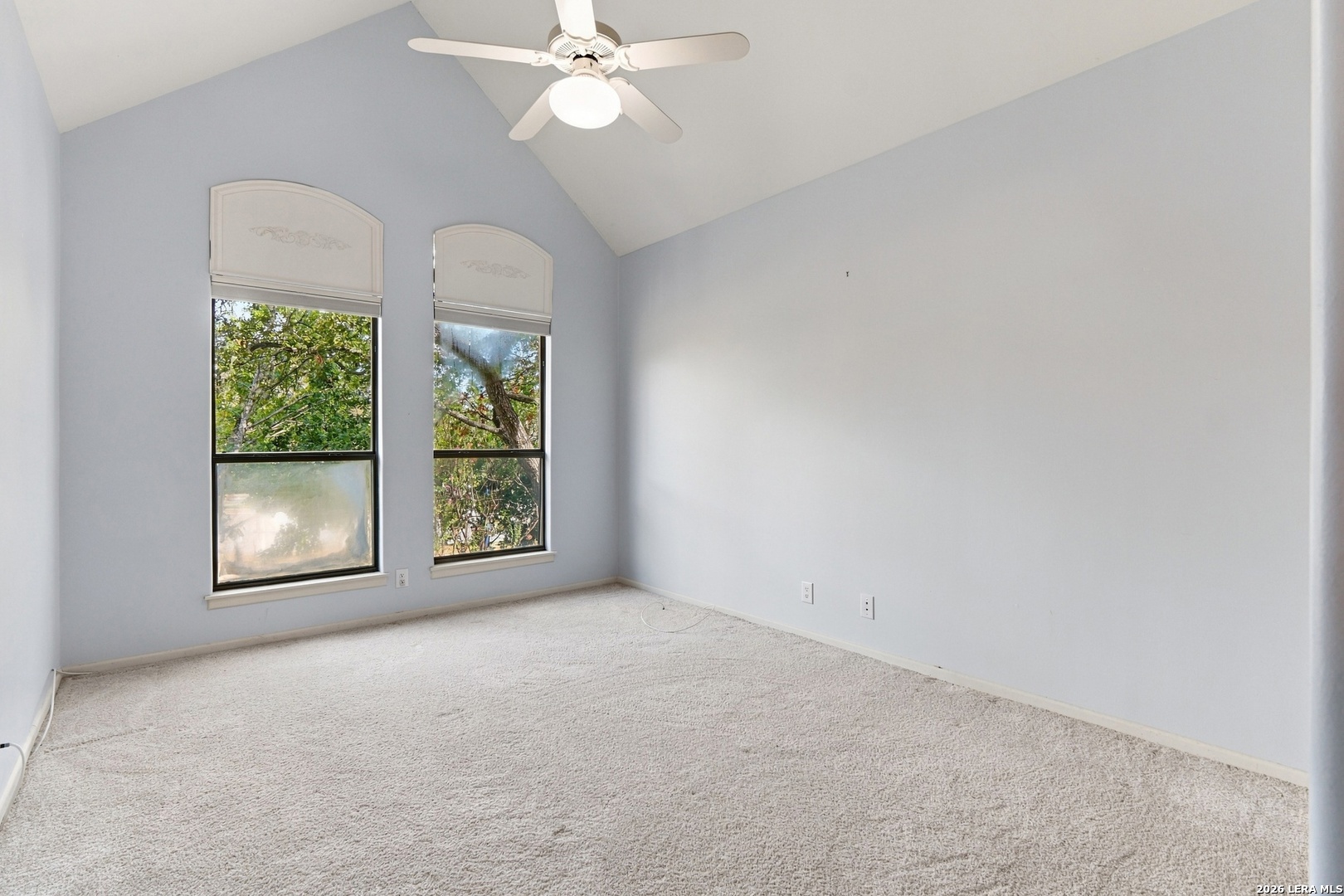 Empty carpeted room with a sloped ceiling, ceiling fan, and two tall windows overlooking trees.