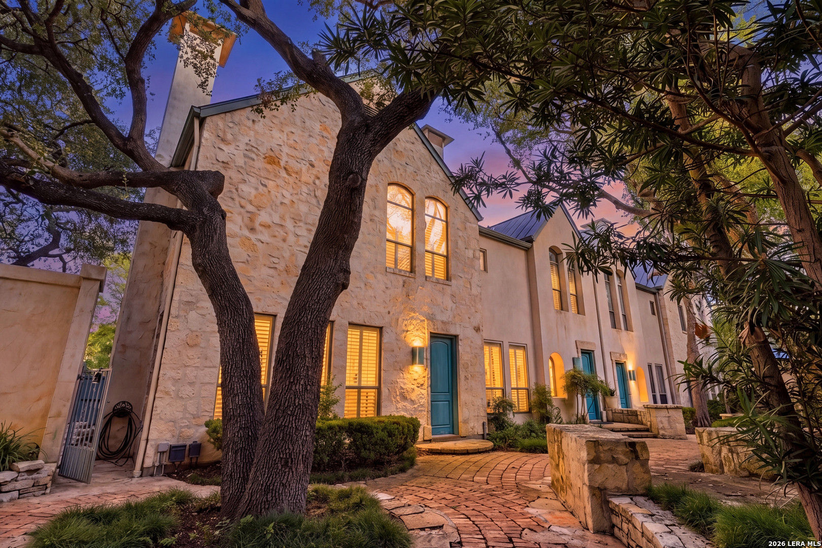 Stone homes with teal doors and glowing windows along a brick walkway beneath mature trees at dusk.