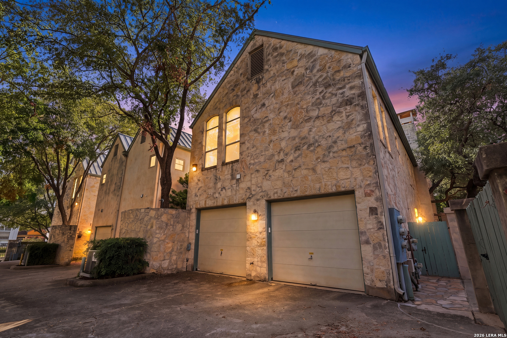 Stone residence at dusk with two garage doors, illuminated windows, and a gated side walkway.