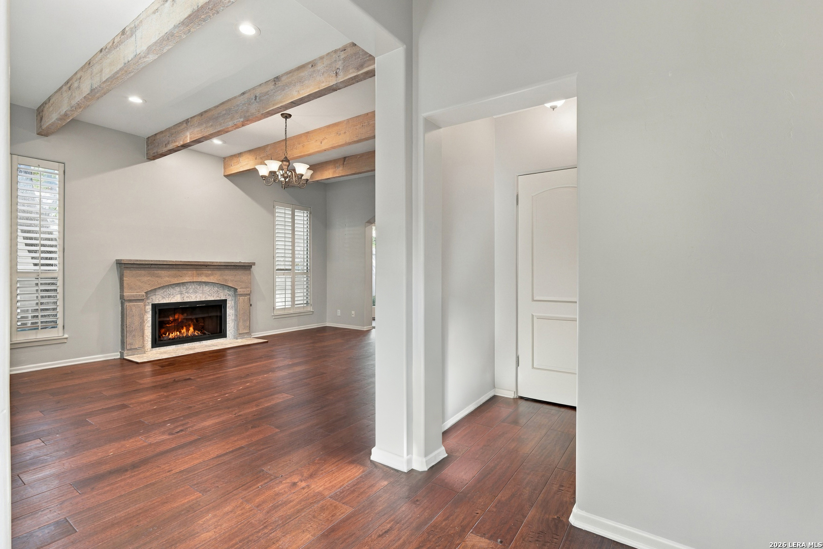 Empty living room with exposed wood beams, dark floors, and a lit stone-surround fireplace.