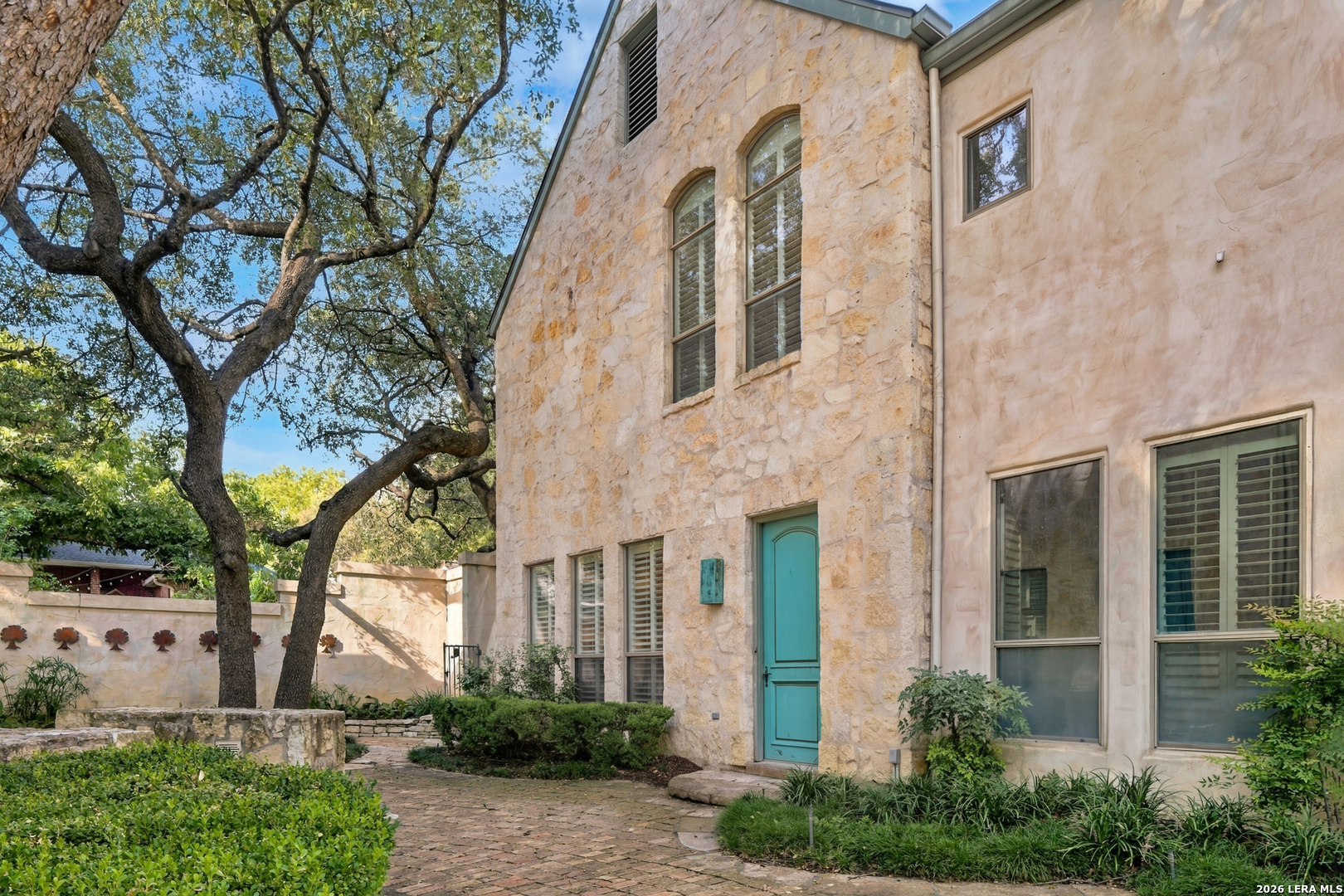 Stone house exterior with turquoise entry door, brick courtyard path, and mature oak trees.