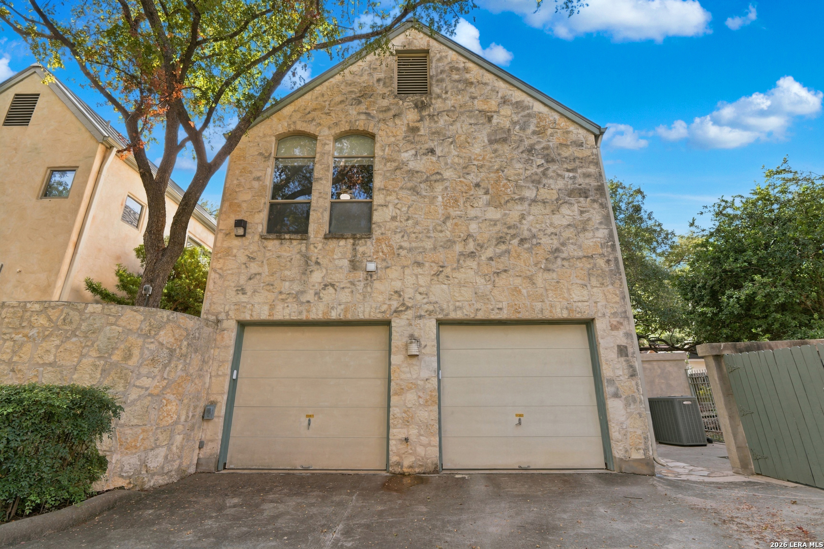 Stone house with two garage doors and arched windows, framed by a large tree.