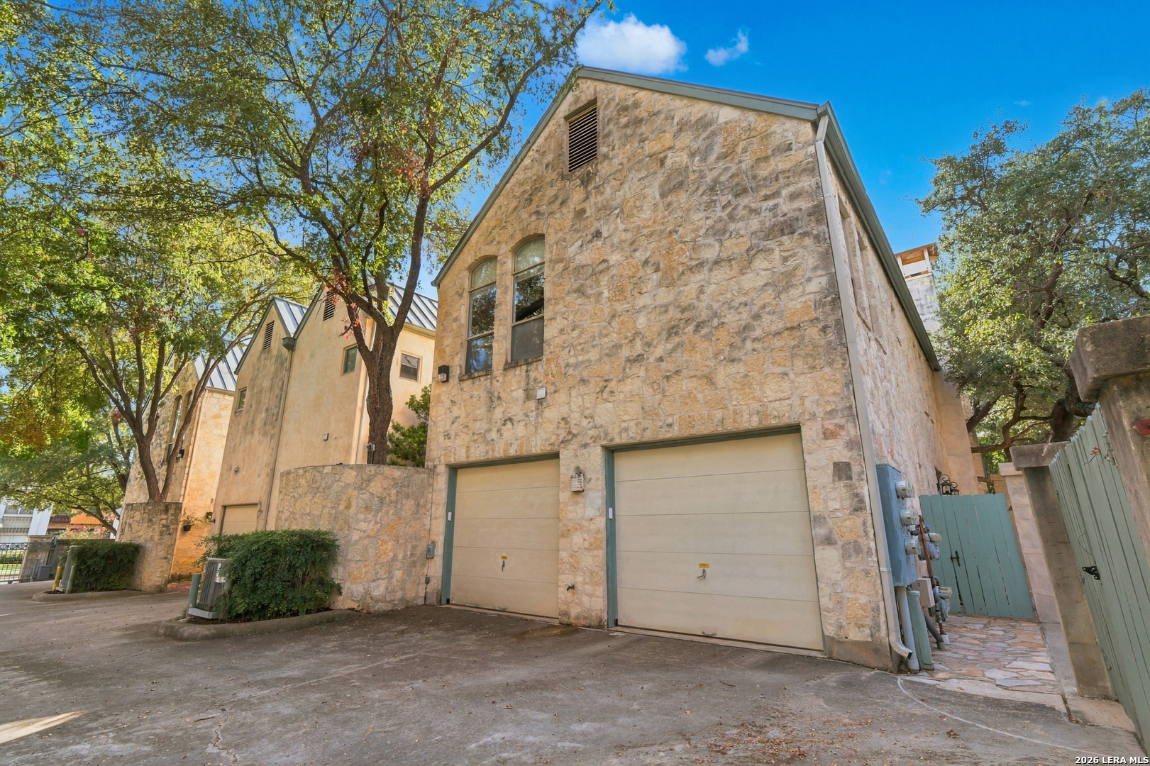 Stone residence with two garage doors and arched windows beneath mature trees.
