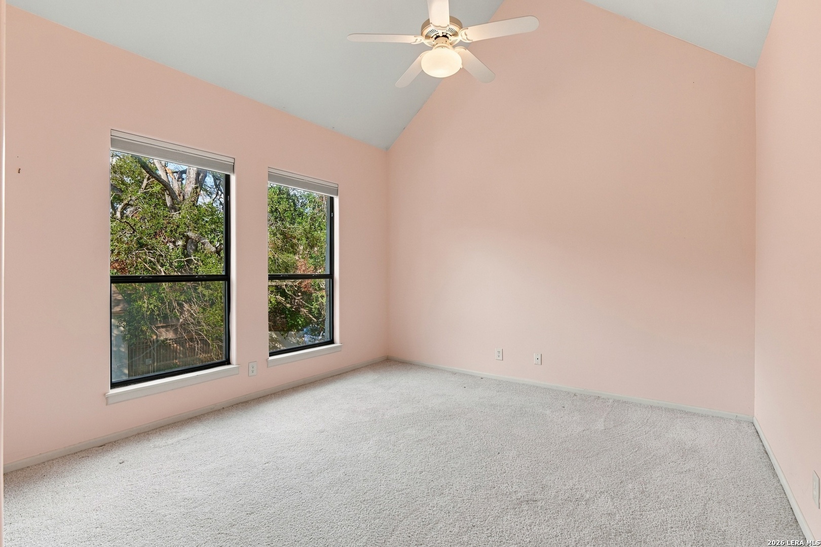 Empty bedroom with pink walls, vaulted ceiling, carpet, and two windows overlooking leafy trees.