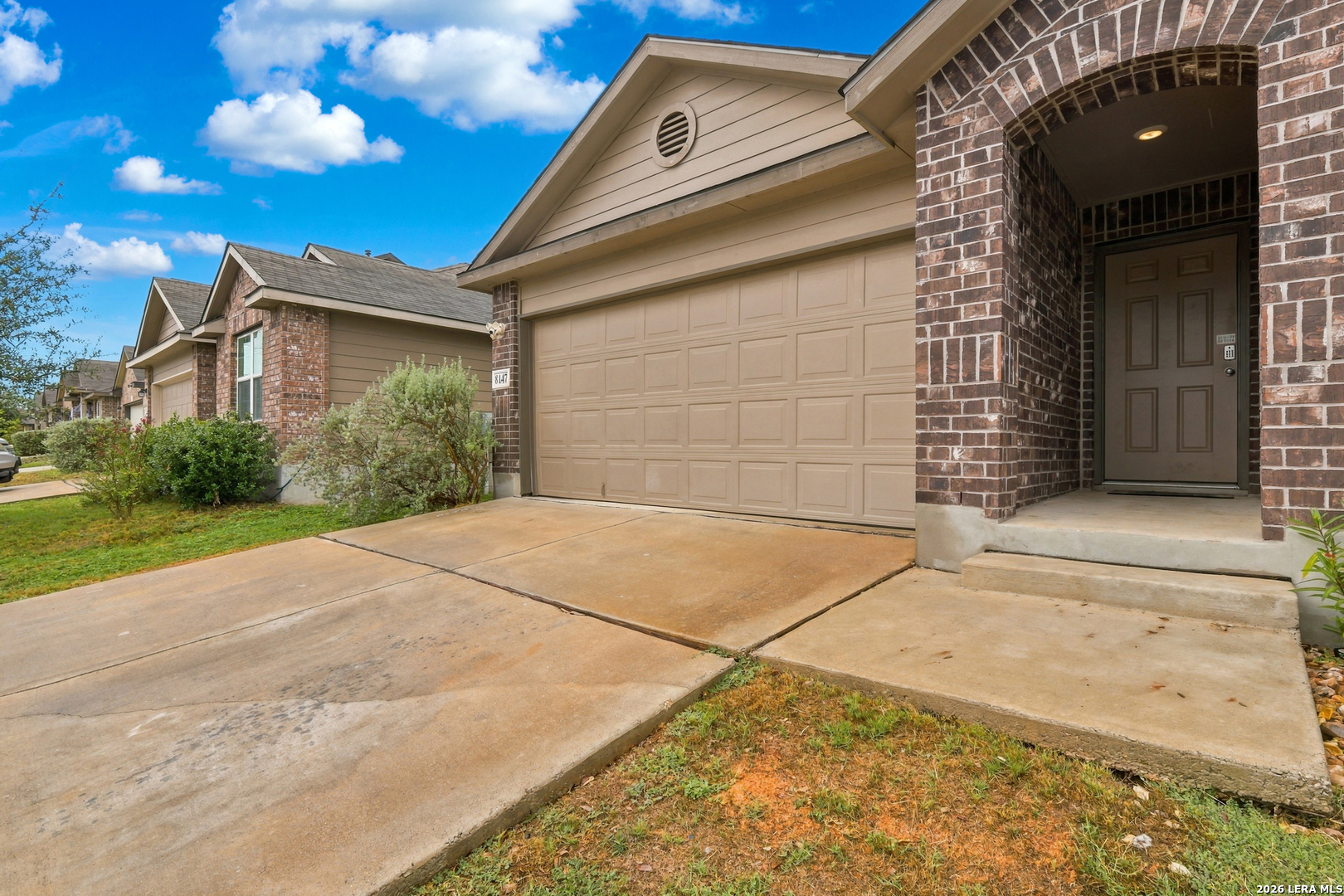 Home exterior with a tan double garage, brick entryway, and broad concrete driveway.