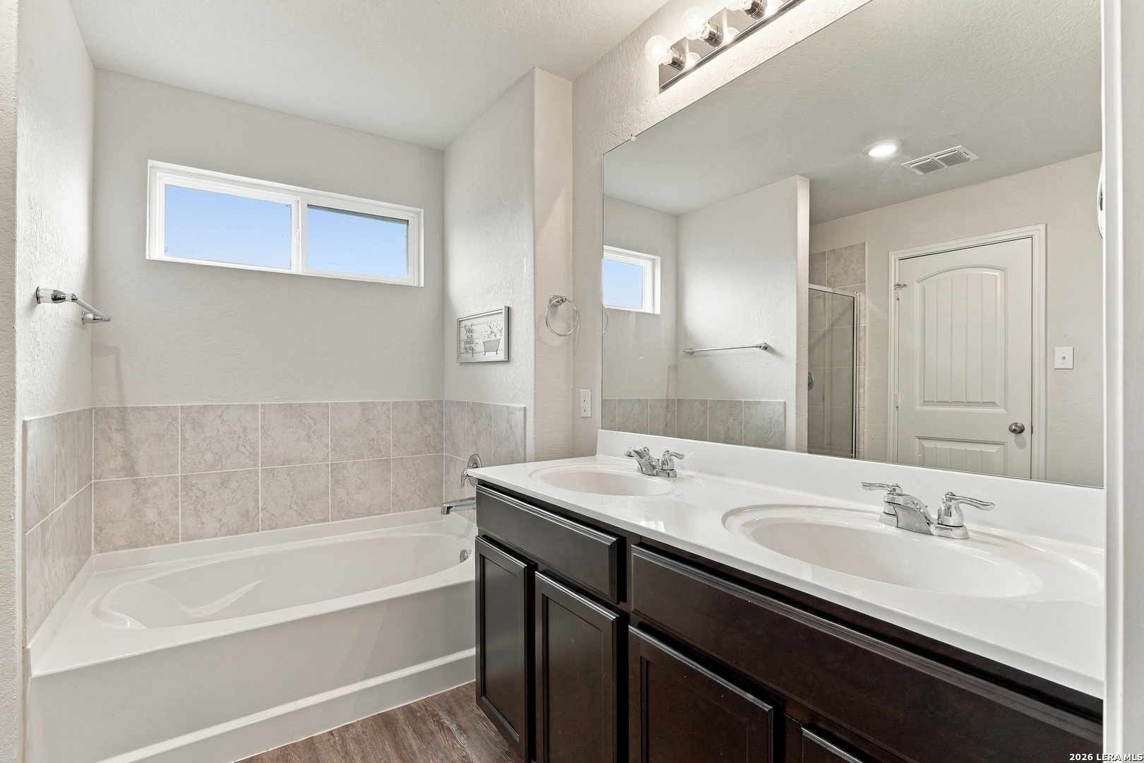 Bathroom with a white tub, beige tile, and a dark double-sink vanity beneath a large mirror.