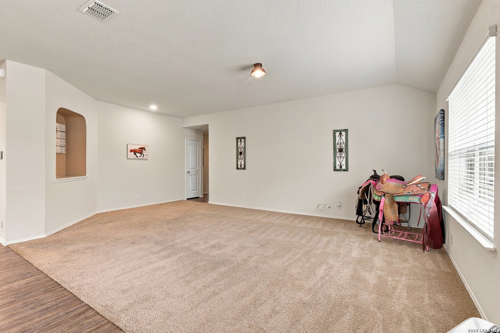 Living room with beige carpet, a large window, and western saddles displayed on stands.