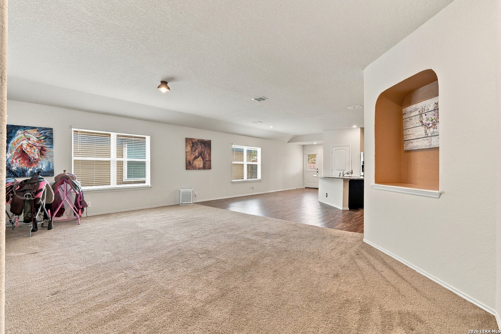 Living room with beige carpet, an open kitchen, and horse saddles beside the windows.