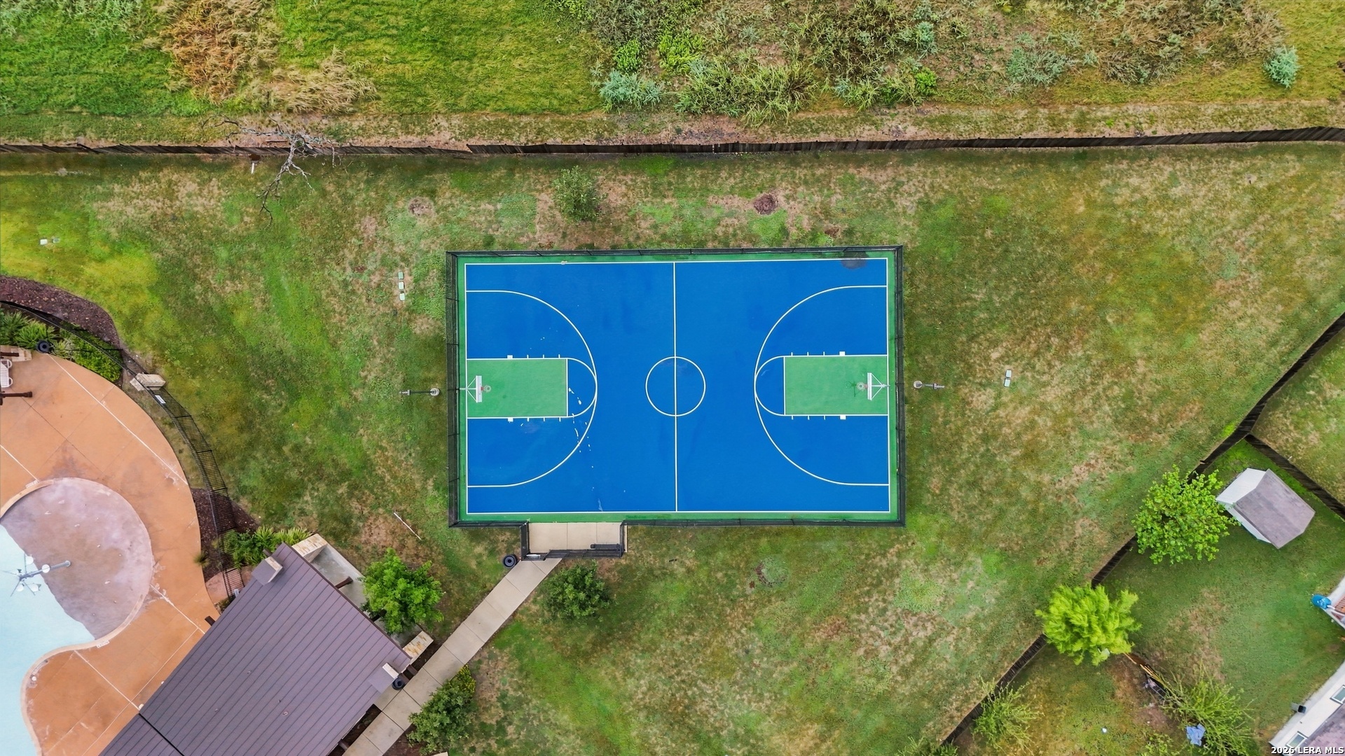 Fenced basketball court in a grassy backyard, beside a swimming pool and covered patio.