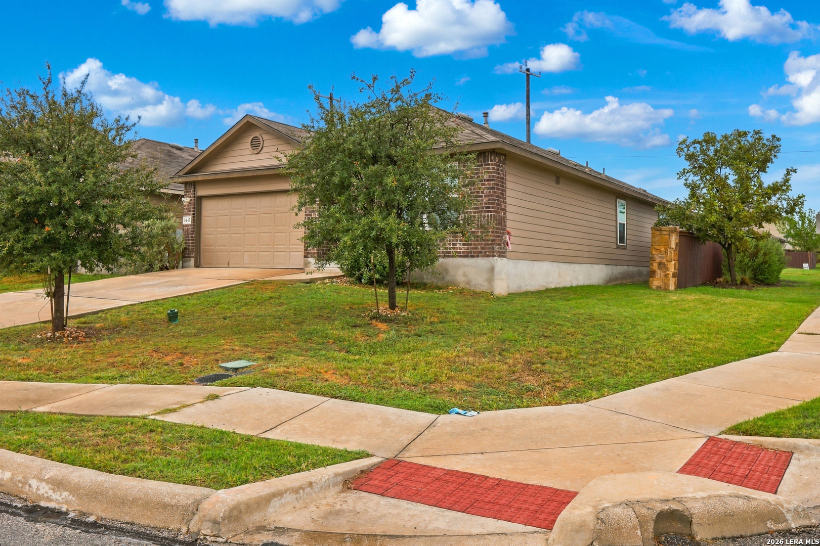 Single-story tan-and-brick house with an attached garage, partly screened by front-yard trees.