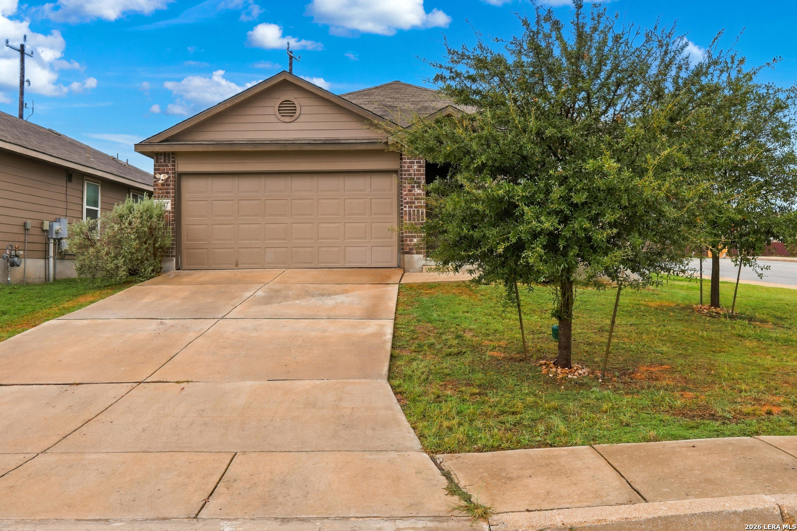 Home with a tan double garage, brick accents, wide driveway, and a tree partly obscuring the front.