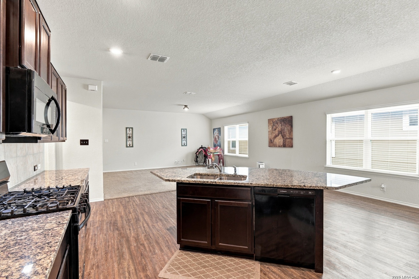 Open-plan kitchen with granite island and dark cabinets, adjoining a bright living area with large windows.