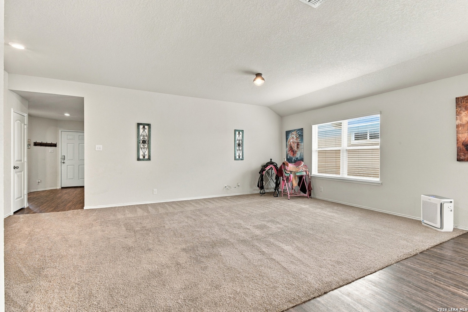 Spacious living room with a large beige rug, window blinds, and saddles displayed beside the window.
