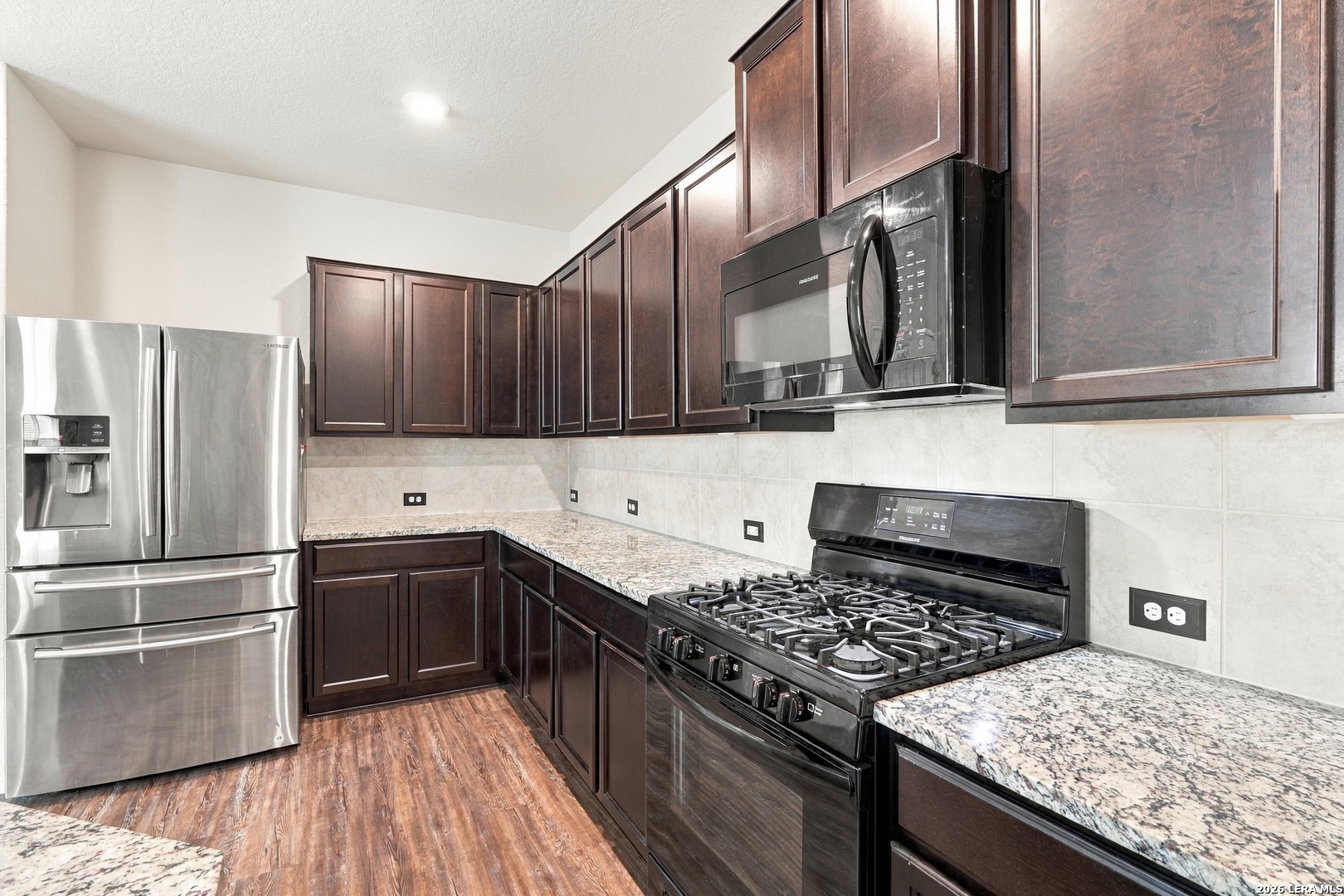 Kitchen with dark wood cabinets, granite counters, stainless refrigerator, and gas range.
