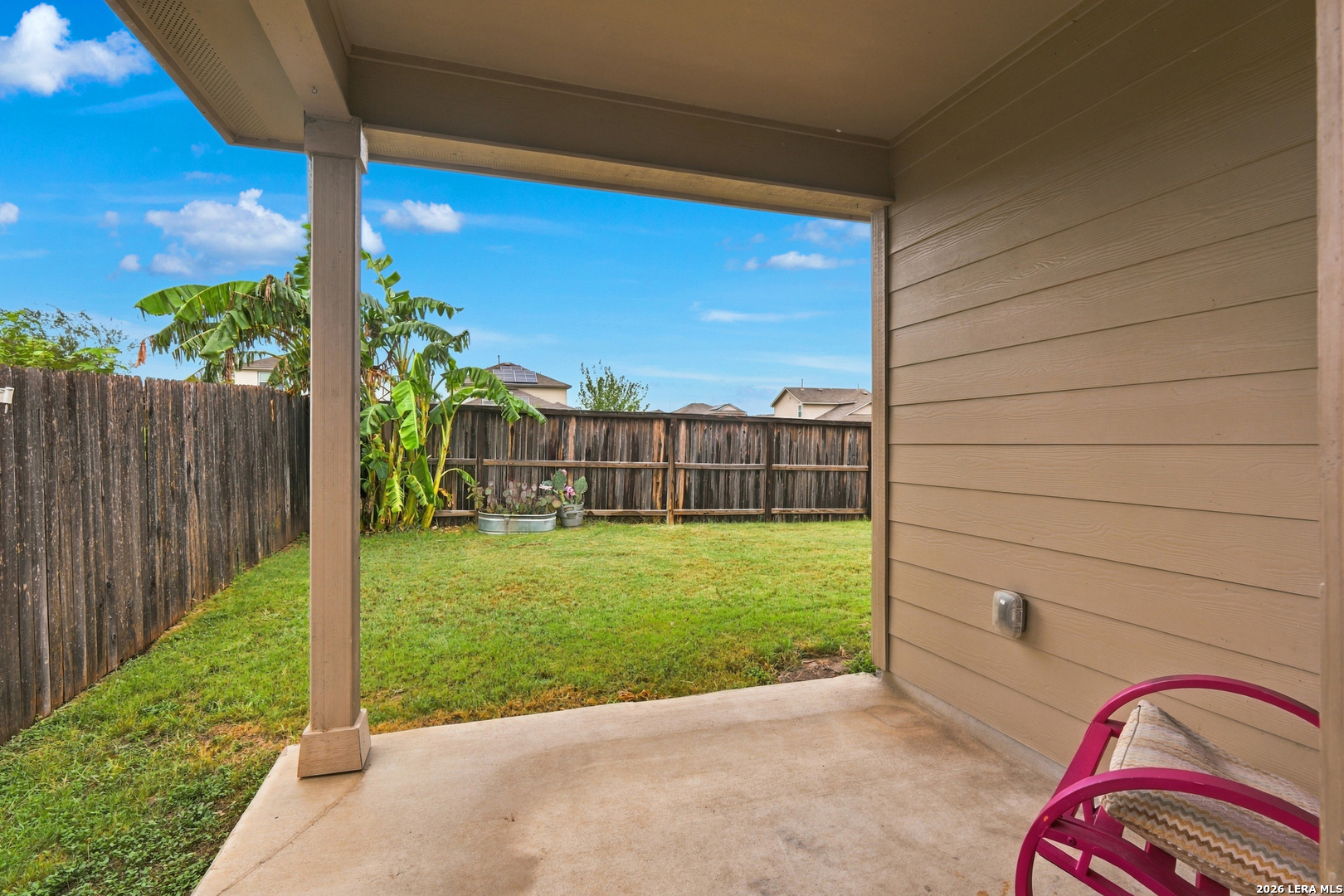 Covered patio overlooking a fenced grassy backyard with banana plants and a pink chair.