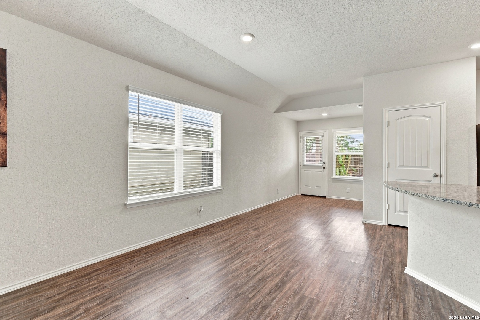 Empty living room with wood-look flooring, high ceiling, large windows, and a granite breakfast bar.