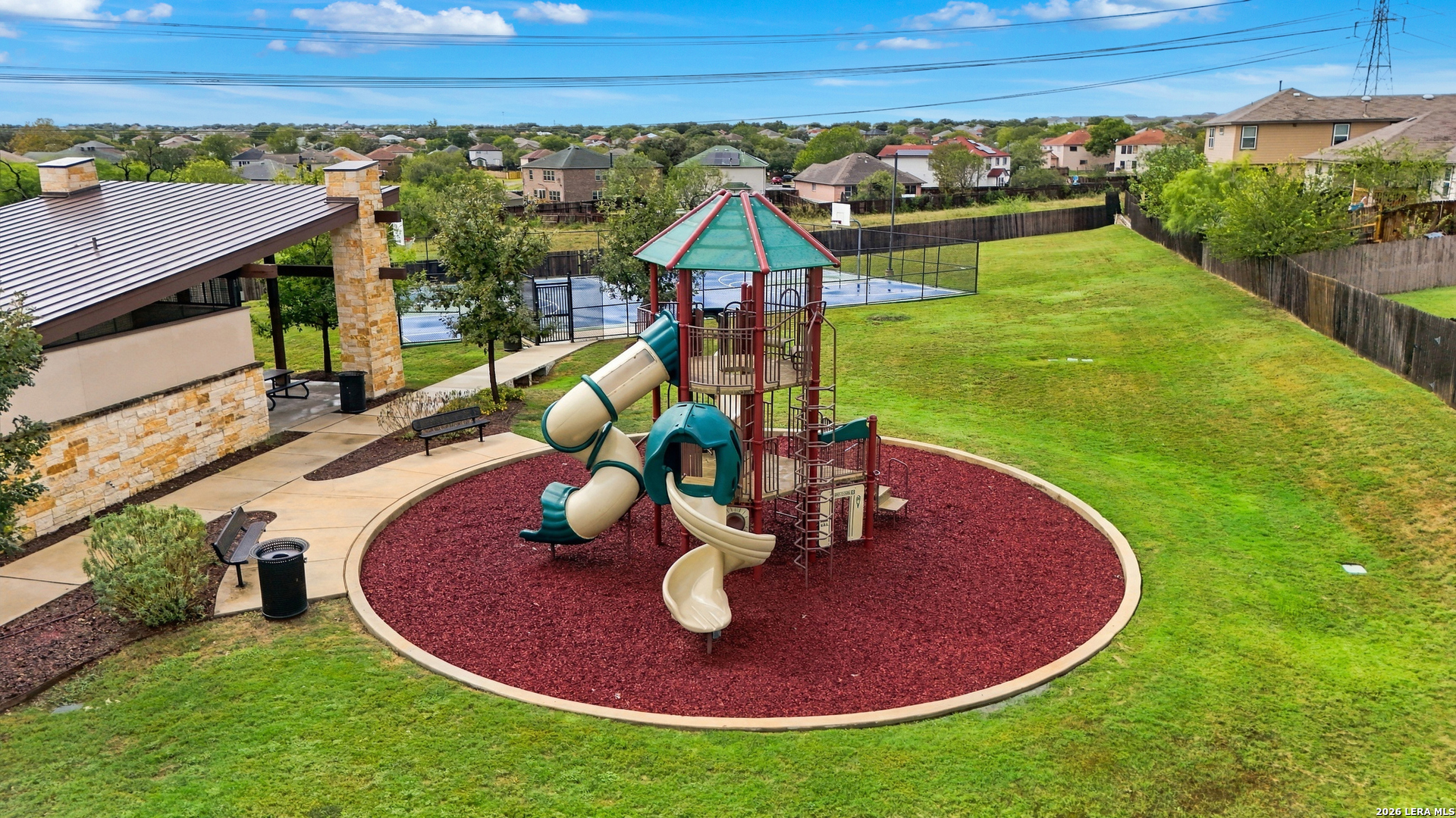 Neighborhood playground with a multilevel play structure, circular mulch area, pavilion, and fenced basketball court.