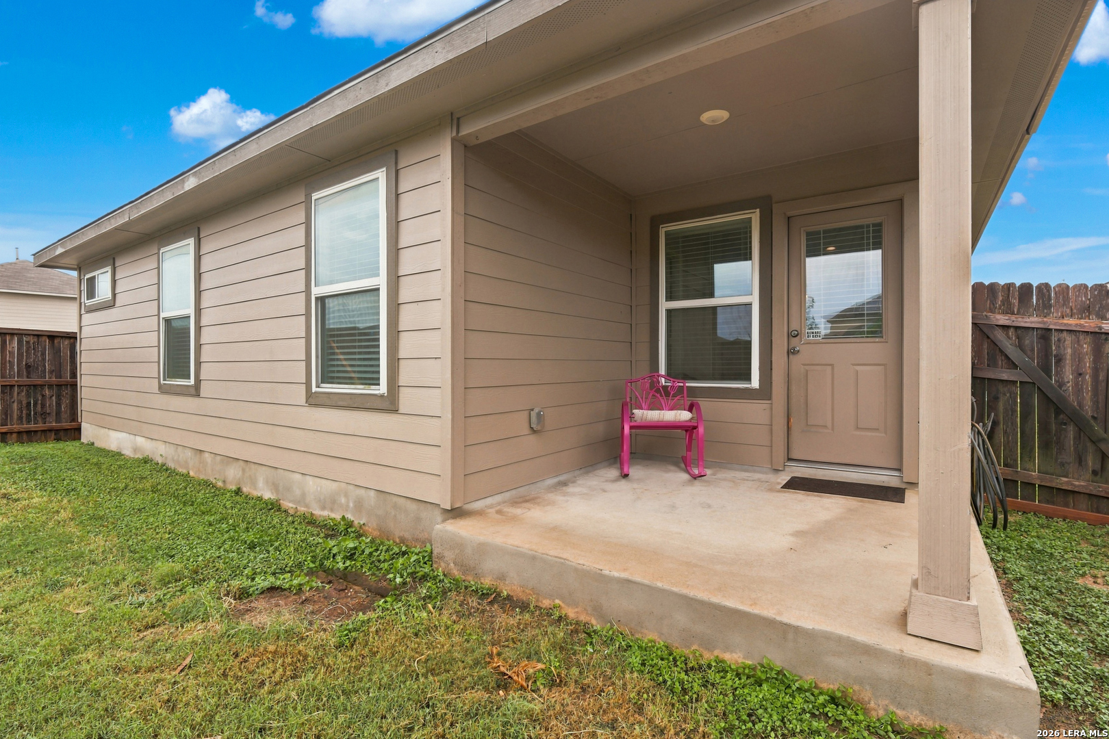 Covered back porch with tan siding, a pink rocking chair, and a fenced grassy yard.