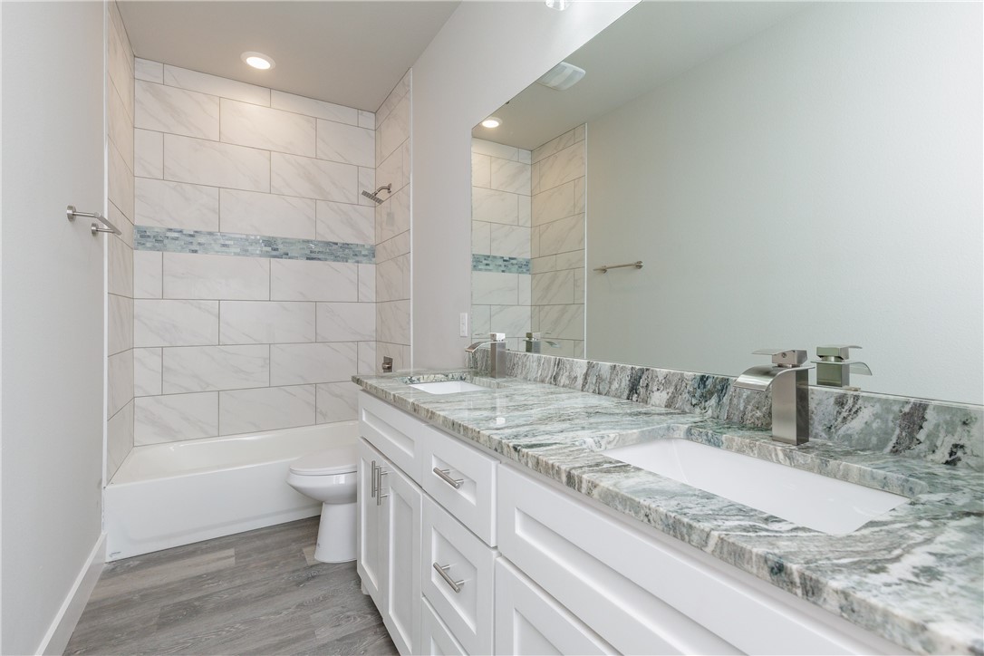 Bathroom with a double vanity, green-gray stone countertop, and marble-look tiled tub-shower surround.