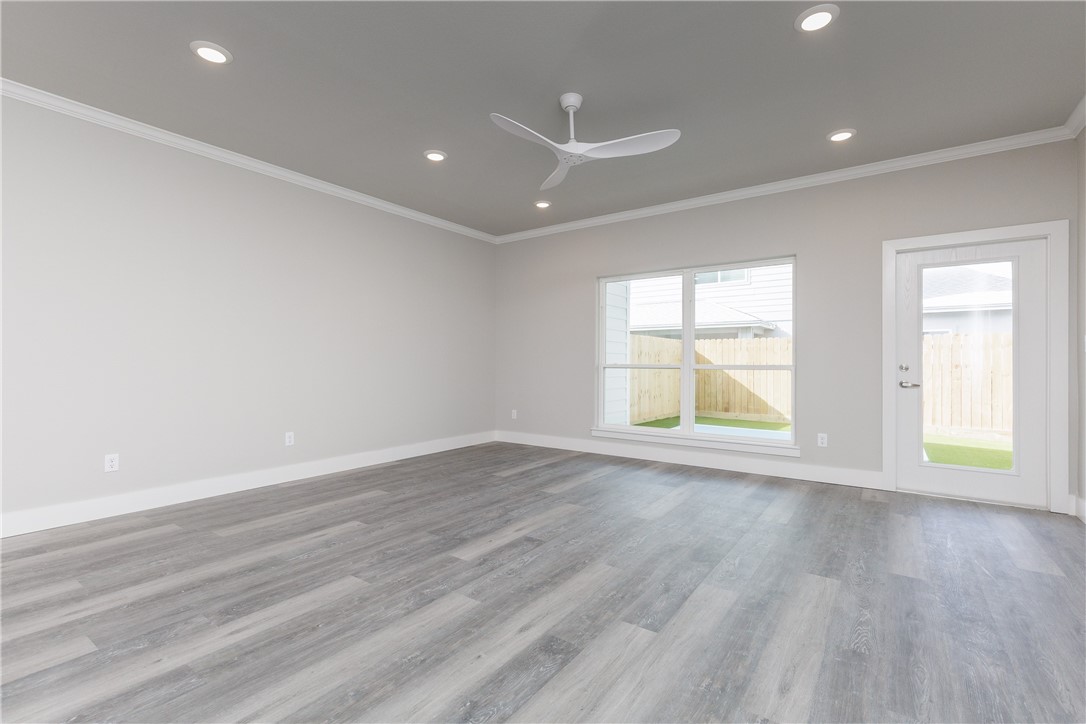 Vacant living room with gray plank flooring, ceiling fan, and windows overlooking a fenced yard.