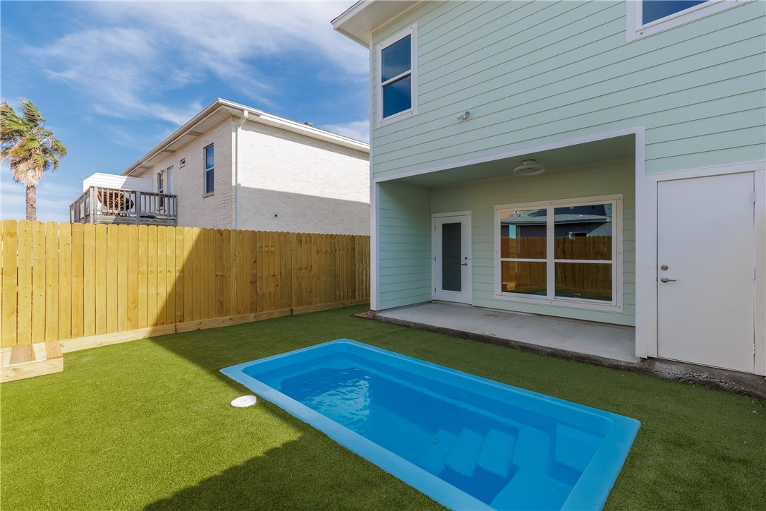 Backyard pool beside a light aqua home, with artificial turf, a wooden fence, and covered patio.