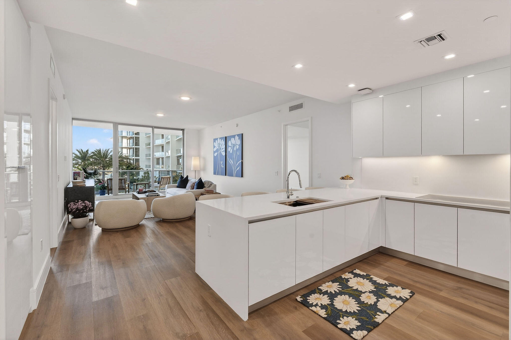 Modern white kitchen with a large island opening to a furnished living area and palm-view windows.