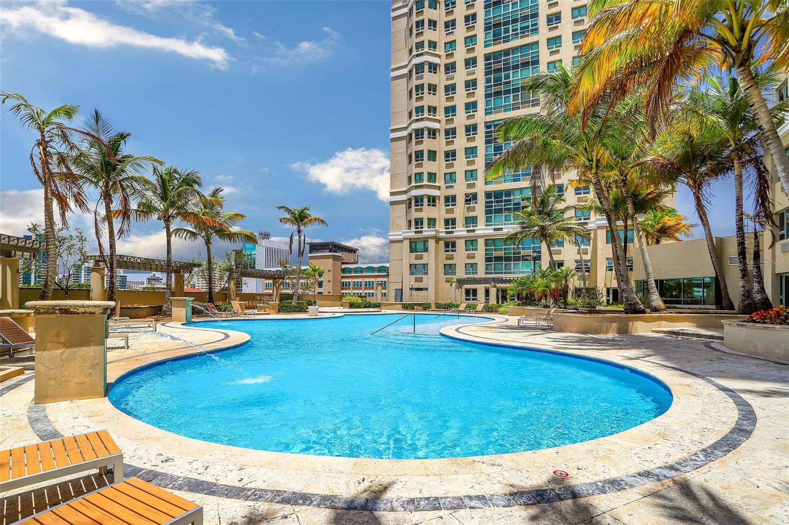 Outdoor condominium pool with a curved stone deck, palm trees, and lounge chairs beside a high-rise tower.