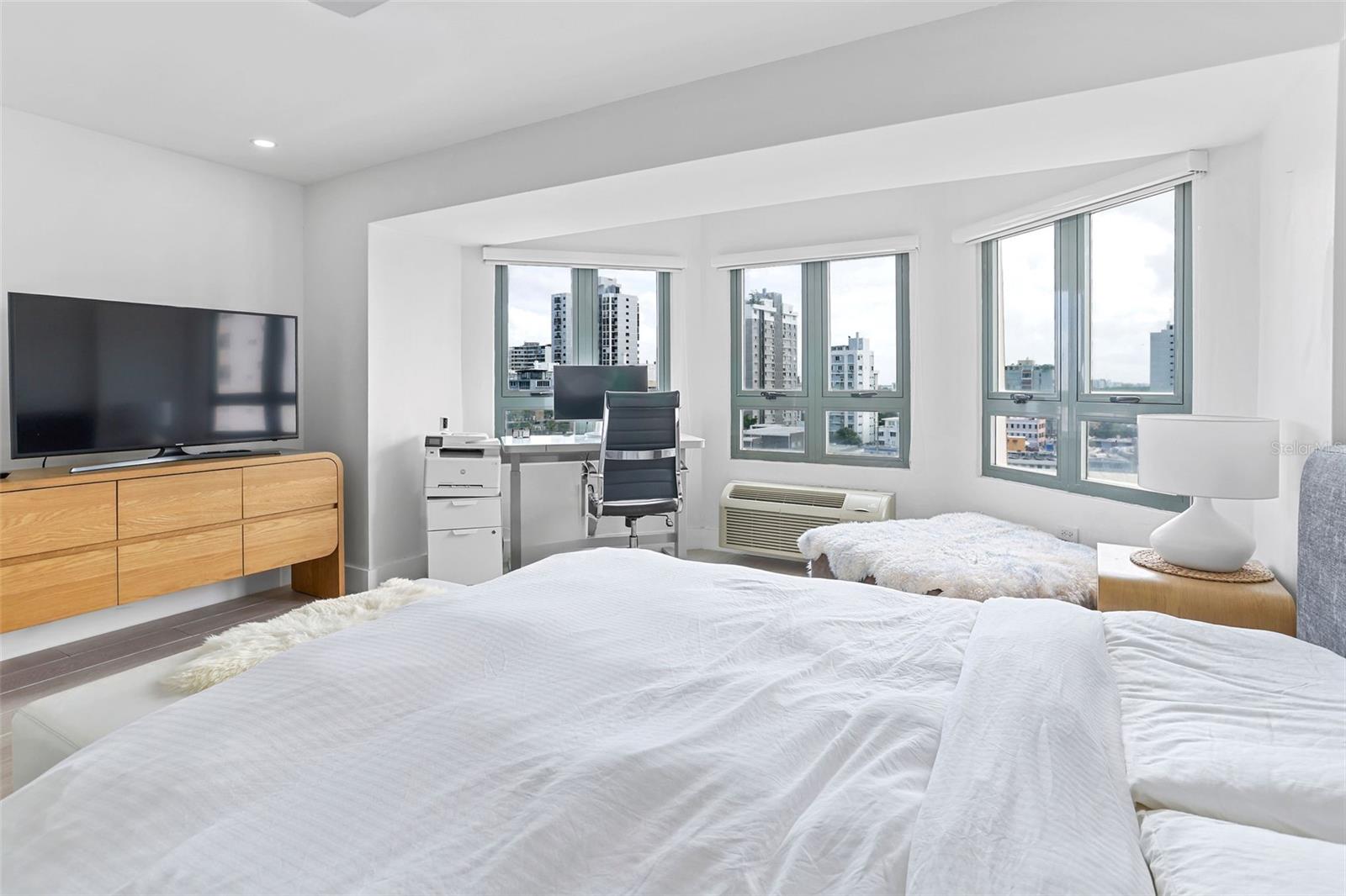 Bedroom with a white bed, desk, and broad windows overlooking city buildings.